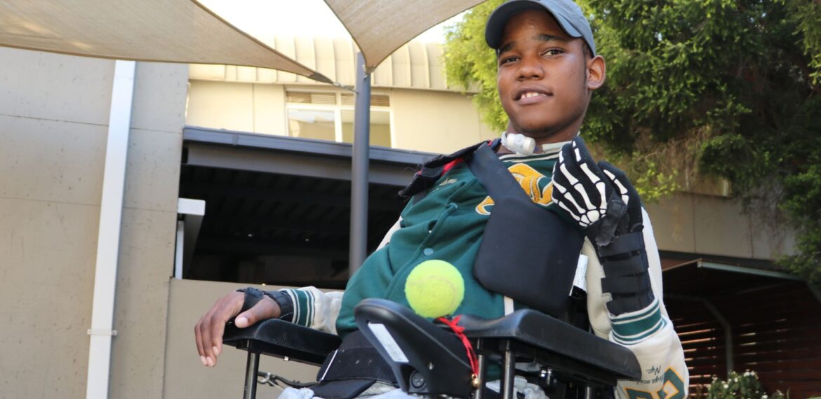 A young man in a wheelchair outdoors wears a cap, sports jacket, and a glove on one hand. He smiles and holds a tennis ball under his arm, with sunshades and a building visible in the background.