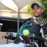 A young man in a wheelchair outdoors wears a cap, sports jacket, and a glove on one hand. He smiles and holds a tennis ball under his arm, with sunshades and a building visible in the background.