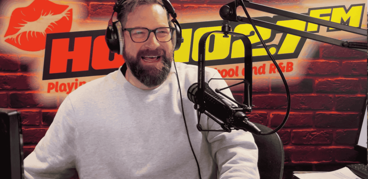 A bearded man wearing glasses and a light gray sweater speaks into a microphone in a radio studio with a red brick wall, HOT 96.7 FM logo, and a red lips graphic in the background. Ian F