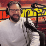 A bearded man wearing glasses and a light gray sweater speaks into a microphone in a radio studio with a red brick wall, HOT 96.7 FM logo, and a red lips graphic in the background. Ian F