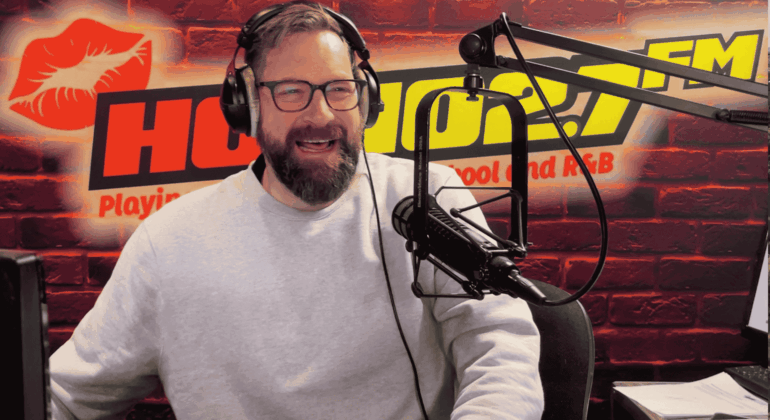 A bearded man wearing glasses and a light gray sweater speaks into a microphone in a radio studio with a red brick wall, HOT 96.7 FM logo, and a red lips graphic in the background. Ian F
