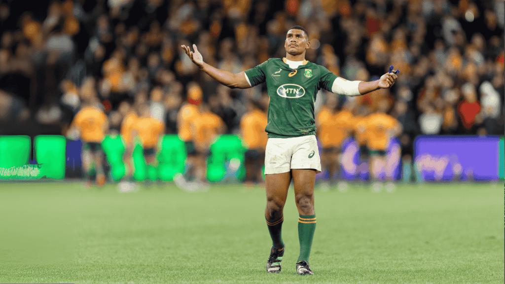A rugby player in a green jersey stands with arms outstretched on a field, looking upward. The background shows blurred players in orange jerseys and a crowd in the stands.
