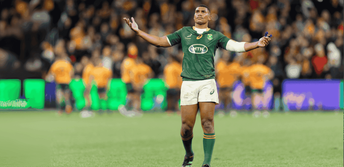 A rugby player in a green jersey stands with arms outstretched on a field, looking upward. The background shows blurred players in orange jerseys and a crowd in the stands.