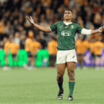 A rugby player in a green jersey stands with arms outstretched on a field, looking upward. The background shows blurred players in orange jerseys and a crowd in the stands.