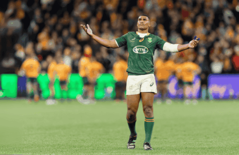 A rugby player in a green jersey stands with arms outstretched on a field, looking upward. The background shows blurred players in orange jerseys and a crowd in the stands.