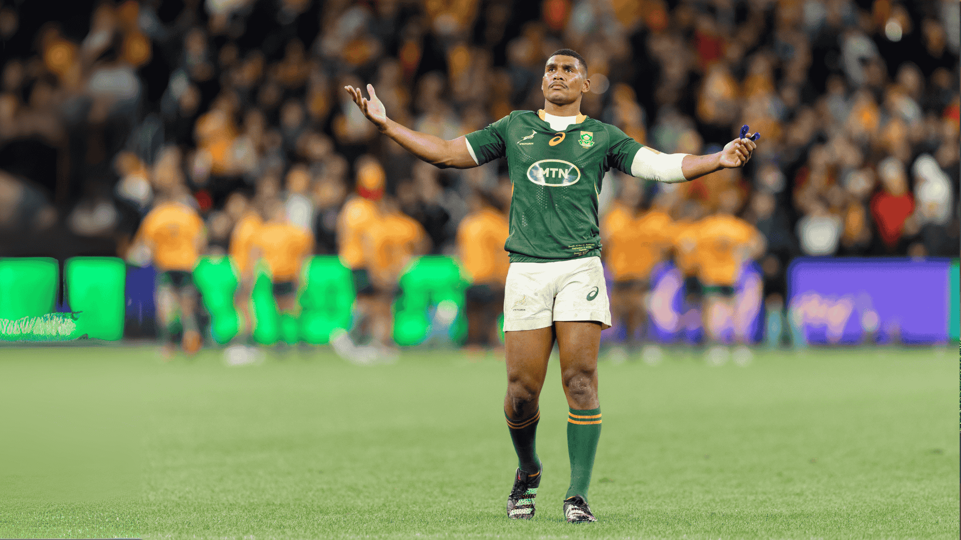 A rugby player in a green jersey stands with arms outstretched on a field, looking upward. The background shows blurred players in orange jerseys and a crowd in the stands.