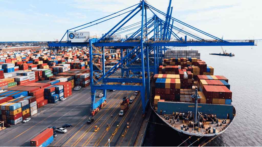 A large cargo ship loaded with colorful shipping containers is docked at the Port of Baltimore, with cranes and stacks of containers visible along the waterfront.