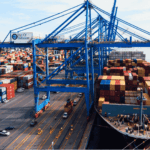 A large cargo ship loaded with colorful shipping containers is docked at the Port of Baltimore, with cranes and stacks of containers visible along the waterfront.