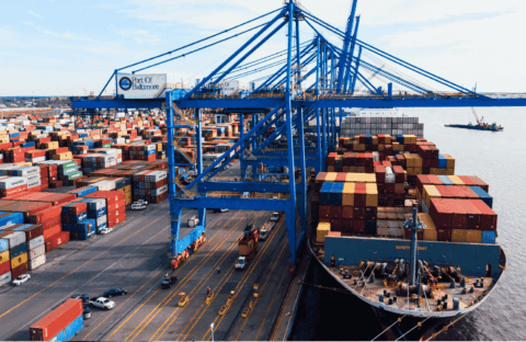 A large cargo ship loaded with colorful shipping containers is docked at the Port of Baltimore, with cranes and stacks of containers visible along the waterfront.