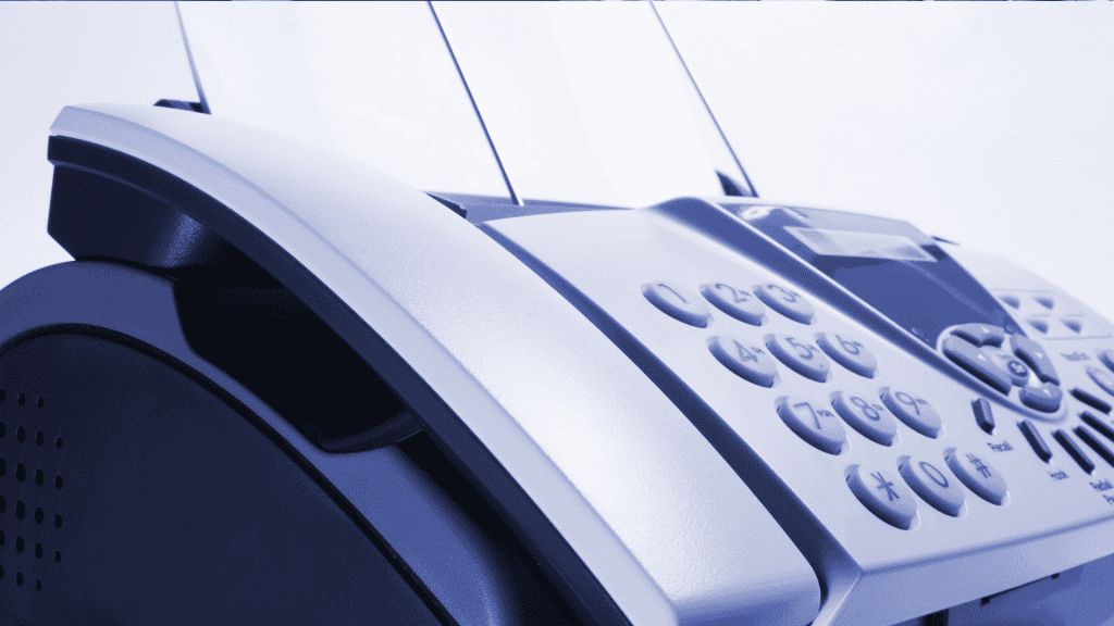 Close-up of a fax machine with paper inserted, showing its keypad and control panel. The image has a blue tint and focuses on the device’s buttons and curved edges.