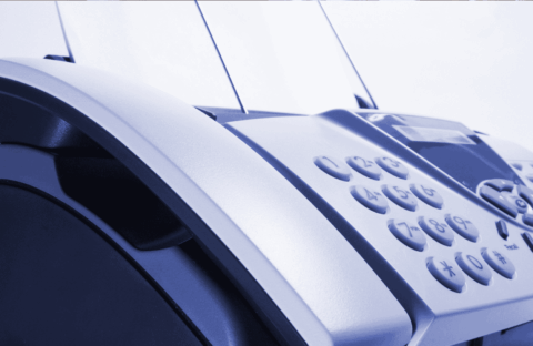 Close-up of a fax machine with paper inserted, showing its keypad and control panel. The image has a blue tint and focuses on the device’s buttons and curved edges.