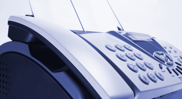 Close-up of a fax machine with paper inserted, showing its keypad and control panel. The image has a blue tint and focuses on the device’s buttons and curved edges.