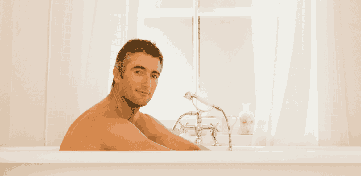 A man with short dark hair sits relaxed in a white bathtub, looking at the camera. The bathroom is bright with white curtains and a classic-style faucet next to him.