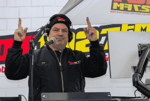 A radio host wearing headphones and a black jacket stands at a broadcast booth, holding both index fingers up. There are microphones, a monitor, and a large 102.7 FM station banner in the background.