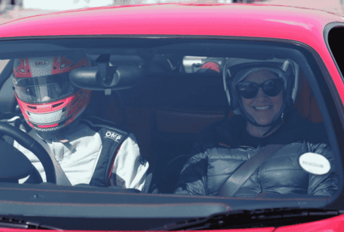 Two people sit in the front seats of a red sports car. The driver wears a racing helmet and suit, while the passenger, smiling, wears a helmet and sunglasses. Both are buckled up, seen through the windshield.