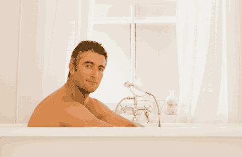 A man with short dark hair sits relaxed in a white bathtub, looking at the camera. The bathroom is bright with white curtains and a classic-style faucet next to him.