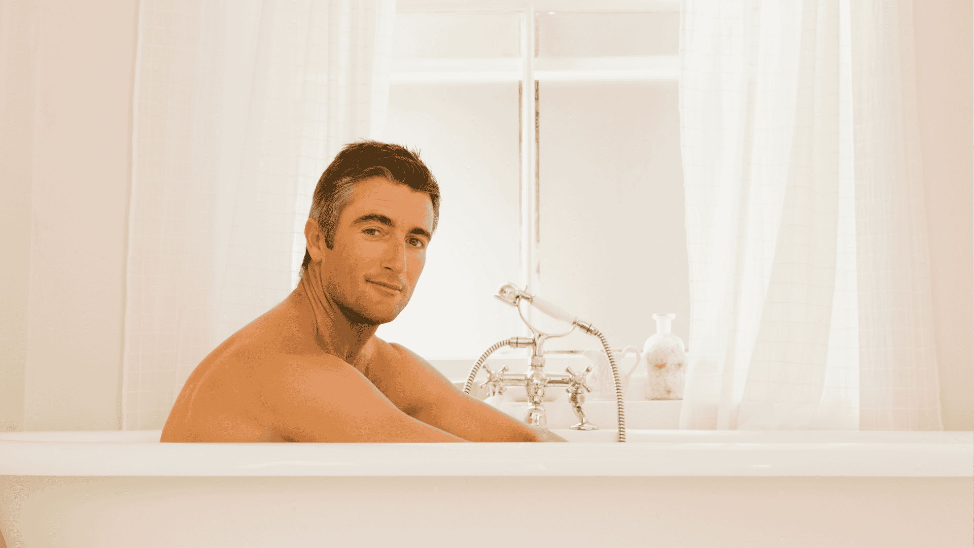 A man with short dark hair sits relaxed in a white bathtub, looking at the camera. The bathroom is bright with white curtains and a classic-style faucet next to him.