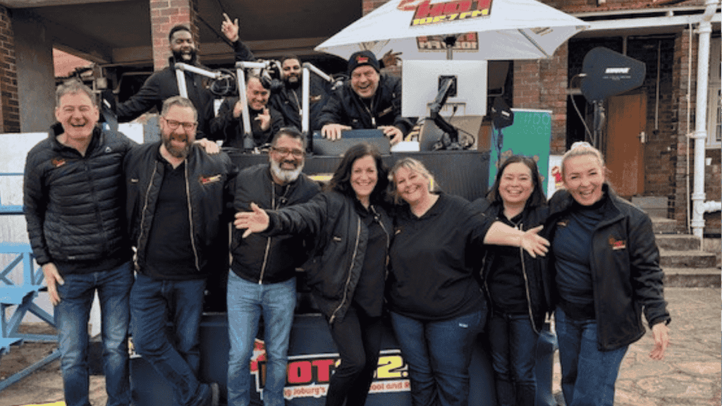 A group of people wearing black jackets stand and smile in front of an outdoor radio station setup, with some people sitting or standing behind a DJ booth under a white umbrella. Everyone looks happy and cheerful.