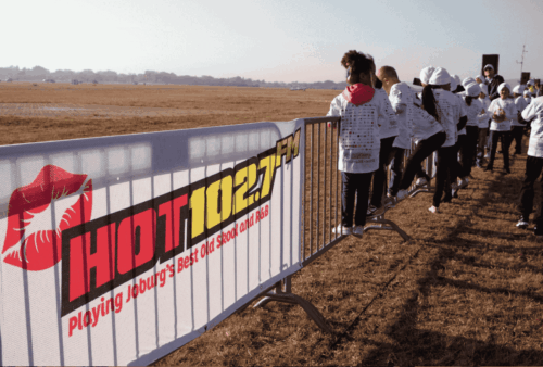 A group of people in matching white jackets walk past a metal barricade with a HOT 102.7 FM banner that reads, “Playing Joburg’s Best Old Skool and R&B,” on a grassy open field.
