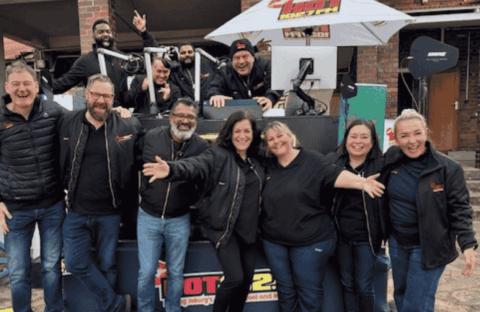 A group of people wearing black jackets stand and smile in front of an outdoor radio station setup, with some people sitting or standing behind a DJ booth under a white umbrella. Everyone looks happy and cheerful.