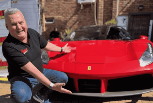 A man in a black polo shirt and jeans kneels and gestures excitedly toward a bright red Ferrari parked on a sunny brick driveway, with banners and a brick wall in the background.