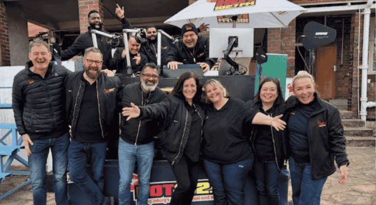 A group of people wearing black jackets stand and smile in front of an outdoor radio station setup, with some people sitting or standing behind a DJ booth under a white umbrella. Everyone looks happy and cheerful.