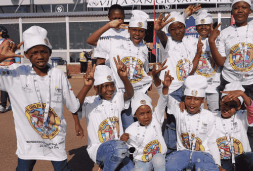 A group of children wearing white chef hats and matching t-shirts pose and smile excitedly outdoors, raising their arms and making playful gestures. A building with signage is visible in the background.