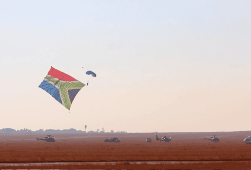 A skydiver descends with a large, colorful South African flag parachute over a flat, open field, with several helicopters and vehicles on the ground below.