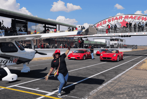 A woman poses and smiles in front of a small airplane and three red sports cars on a racetrack. Spectators watch from the stands under the Red Star Raceway arch on a sunny day.