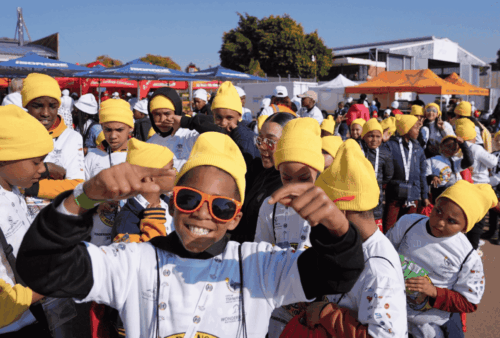 A large group of smiling children wearing yellow beanies and sunglasses gather outdoors at an event, holding medals and dressed in matching white shirts with logos. Colorful tents and other people are visible in the background.