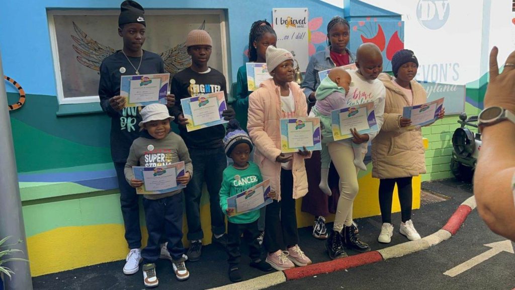 A group of children and teens stand outdoors, holding certificates and smiling for a photo. They are dressed warmly in jackets and hats, with colorful walls behind them. An adult’s hand is visible, taking the picture.