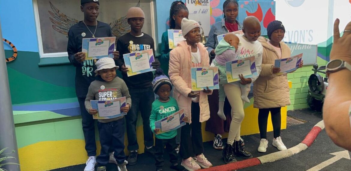 A group of children and teens stand outdoors, holding certificates and smiling for a photo. They are dressed warmly in jackets and hats, with colorful walls behind them. An adult’s hand is visible, taking the picture.