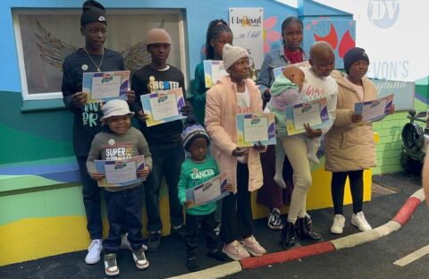 A group of children and teens stand outdoors, holding certificates and smiling for a photo. They are dressed warmly in jackets and hats, with colorful walls behind them. An adult’s hand is visible, taking the picture.