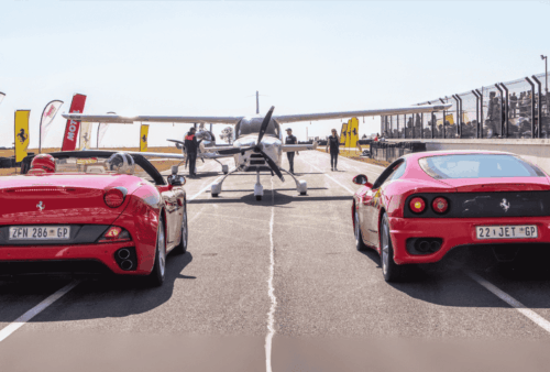 Two red Ferraris are lined up on a racetrack next to a small aircraft, with people and banners in the background, suggesting a race or automotive event.