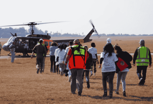 A group of people, some wearing safety vests, walk across an open field toward a helicopter with spinning blades on a sunny day.