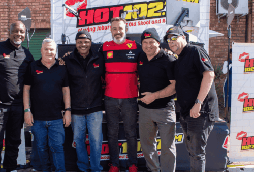 Six men stand smiling in front of a Hot 102.7 FM promotional backdrop, some with arms around each other. Most are wearing black shirts with the station’s logo; one wears a red Ferrari shirt. They are outdoors on a sunny day.