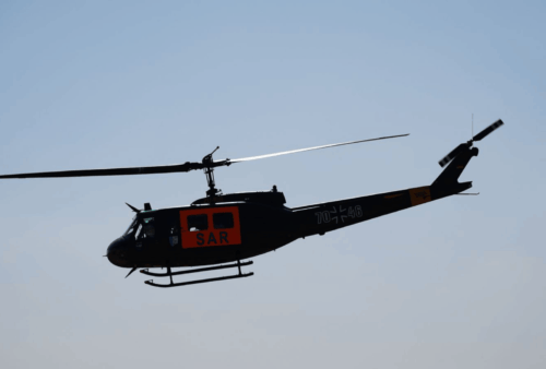 A black helicopter with SAR marked in orange on its side flies against a clear blue sky. The main and tail rotors are visible, and some details and markings are seen on the helicopter’s body.