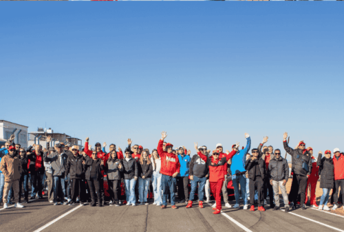 A large group of people standing on a road, many wearing red clothing, waving at the camera under a clear blue sky.