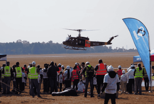 A helicopter hovers above a grassy field while a crowd of people wearing high-visibility vests and helmets watch from behind a rope barrier. Colorful tents and flags are set up nearby. Trees line the background.