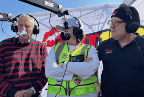 Three people stand together at an outdoor radio broadcast. One man in a striped sweater speaks into a microphone, while a woman in a safety vest and a man in glasses and a beanie stand beside her, each wearing headphones.
