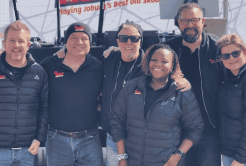 Six people wearing black jackets stand together outdoors, smiling at the camera. Some have their arms around each other. A sign with red text is partially visible in the background.