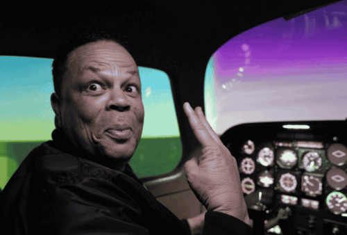 A man sits in an airplane cockpit, smiling playfully and holding up two fingers in a peace sign gesture. The cockpit displays glowing controls and a colorful sky visible through the windows.
