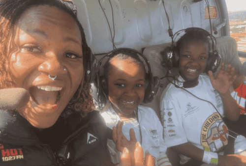 A woman and two children, all smiling and wearing headsets, sit inside a helicopter. The children wear white t-shirts, and sunlight streams in through the windows. Everyone appears excited and happy.