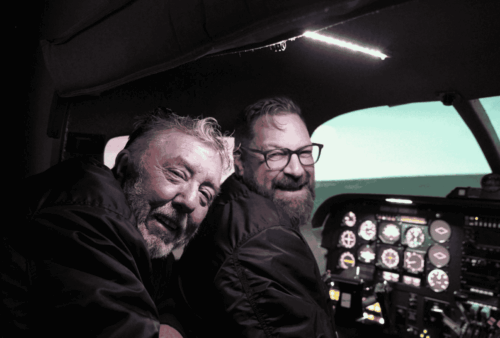 Two men wearing dark jackets sit in an airplane cockpit, smiling at the camera. The cockpit is illuminated, showing various control panels and instruments, with a view of the sky through the windshield.