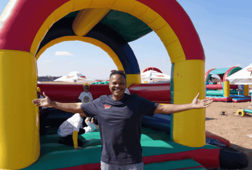 A smiling man stands with arms outstretched in front of a colorful inflatable bounce house at an outdoor event on a sunny day. Children play on the inflatables in the background.