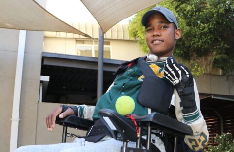 A young man in a wheelchair outdoors wears a cap, sports jacket, and a glove on one hand. He smiles and holds a tennis ball under his arm, with sunshades and a building visible in the background.