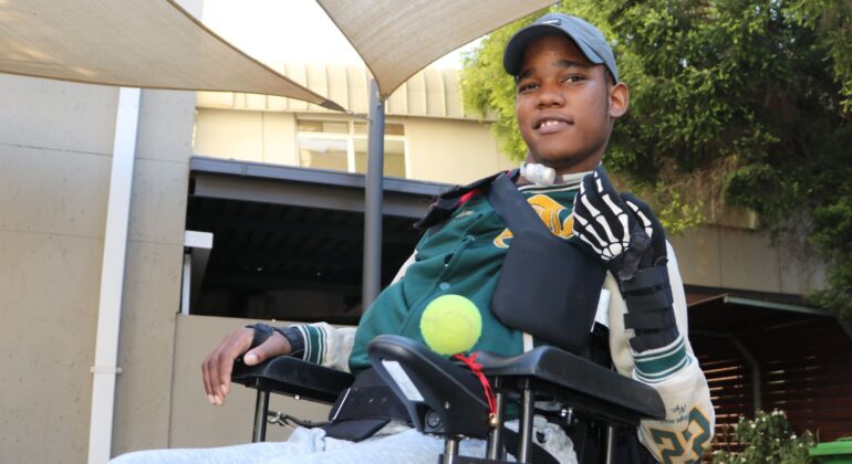 A young man in a wheelchair outdoors wears a cap, sports jacket, and a glove on one hand. He smiles and holds a tennis ball under his arm, with sunshades and a building visible in the background.