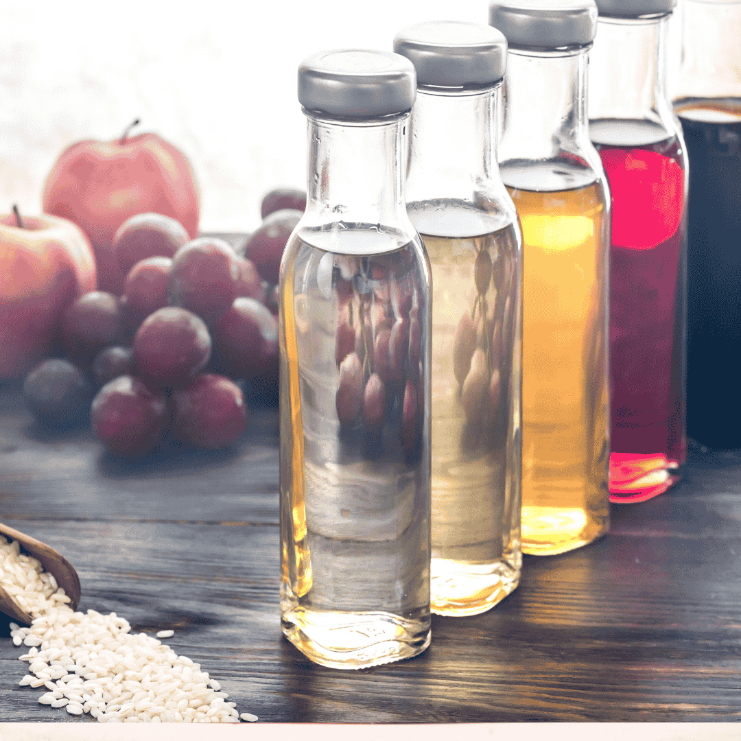 Four glass bottles filled with different types of vinegar are arranged in a row on a wooden surface. Grapes, apples, and loose grains are visible in the background.