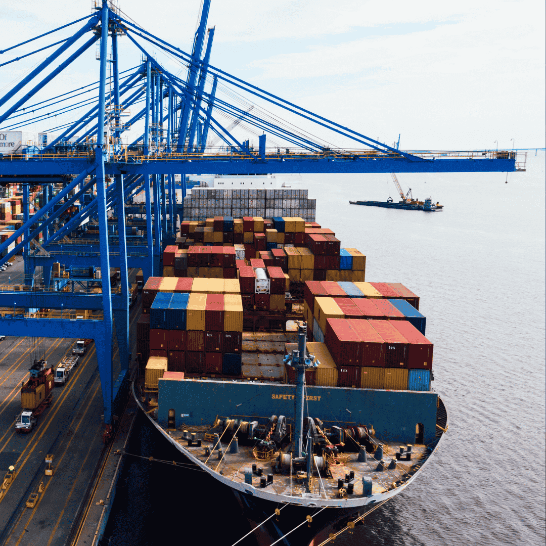 A large cargo ship loaded with colorful shipping containers is docked at a busy port with blue cranes. The sea and another small vessel are visible in the background.