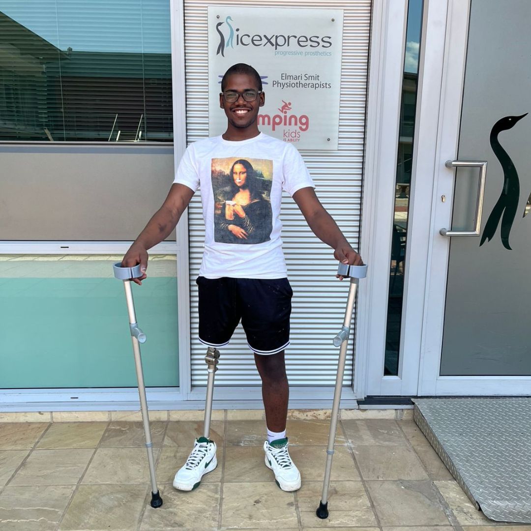 A smiling young man with a leg prosthesis stands with crutches outside a physiotherapy clinic. He wears glasses, a Mona Lisa t-shirt, black shorts, and white sneakers.